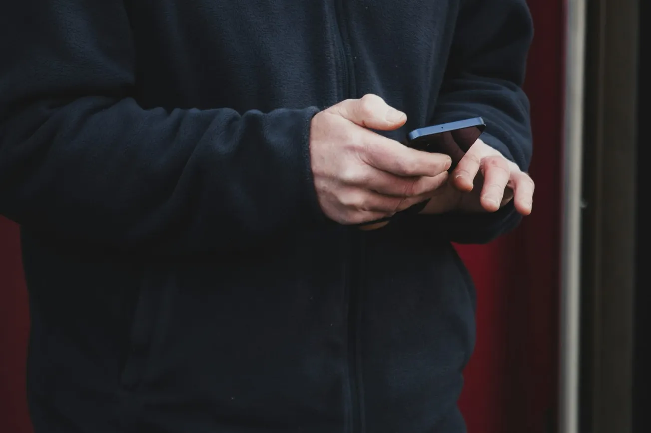 A person in a dark jacket uses a smartphone with both hands. The background is blurred, creating a focused and contemplative mood.