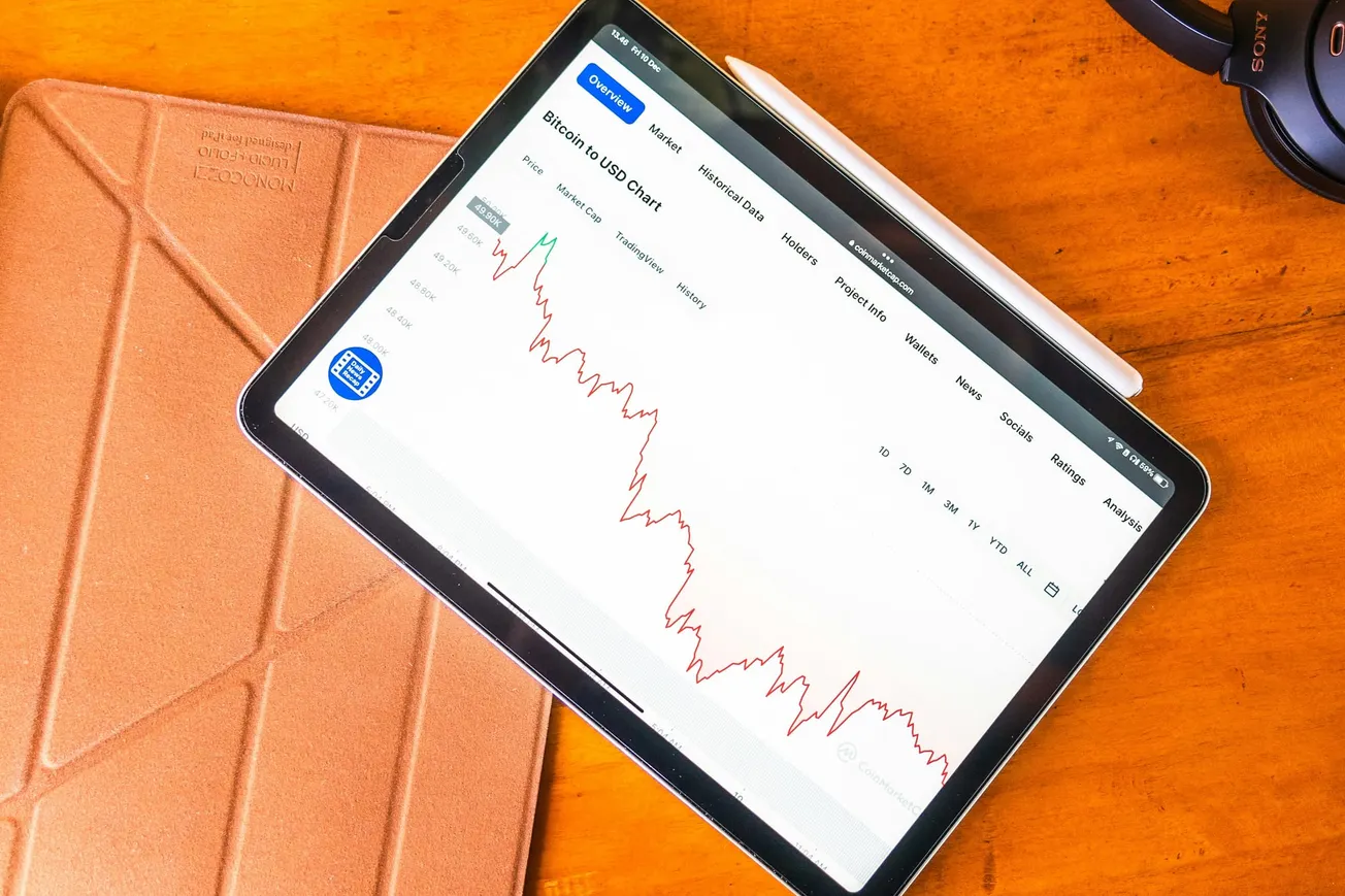 A tablet displays a Bitcoin to USD price chart showing a significant downward trend on a white background, resting on a wooden desk next to a stylus and headphones.