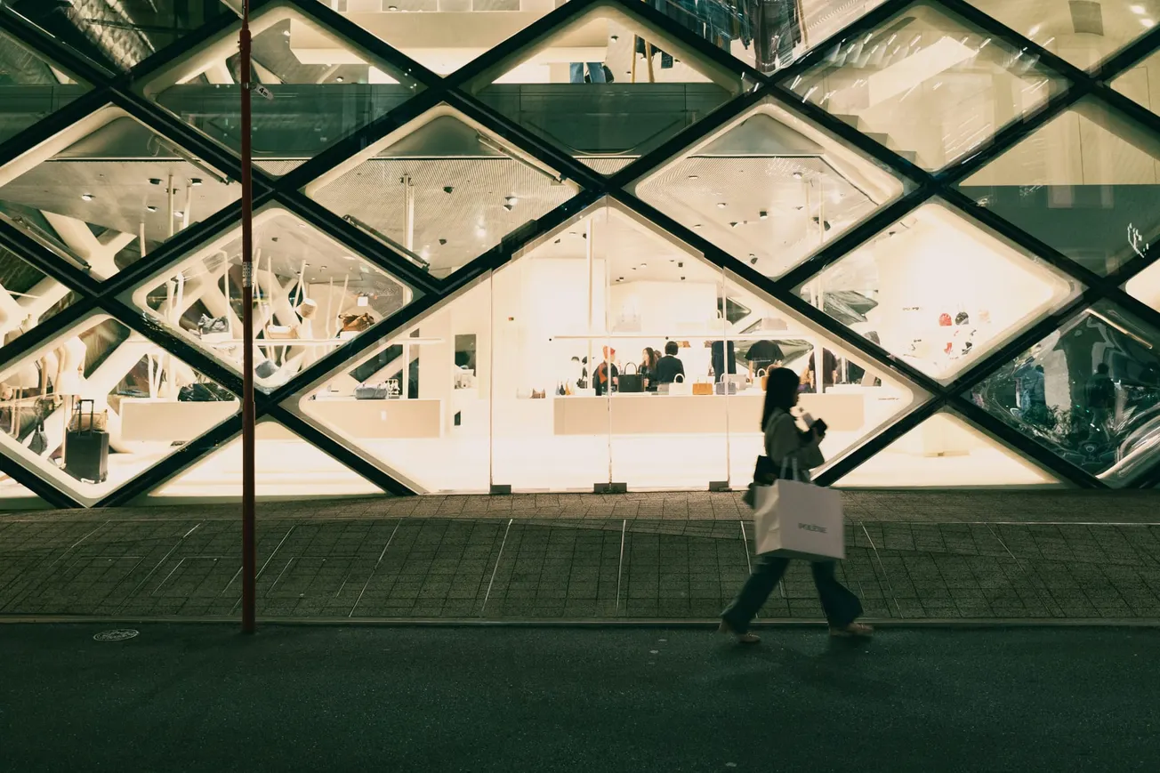 A woman walks past a modern building with diamond-patterned glass. Inside, a bright, minimalistic store is visible. The scene is calm and urban.