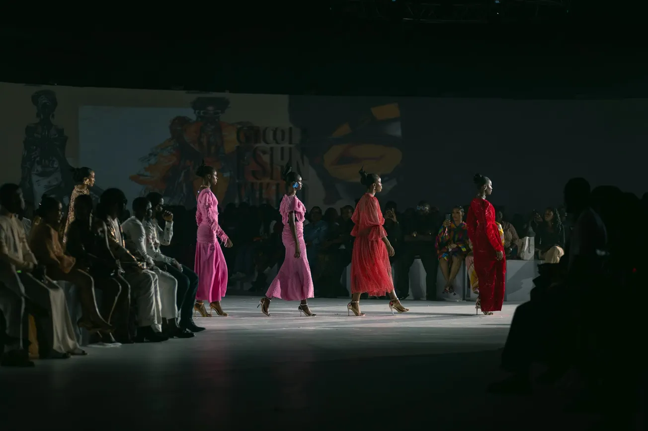 Models walk on a dimly lit runway in vibrant pink and red dresses, watched by an attentive audience, creating an elegant and dynamic atmosphere.