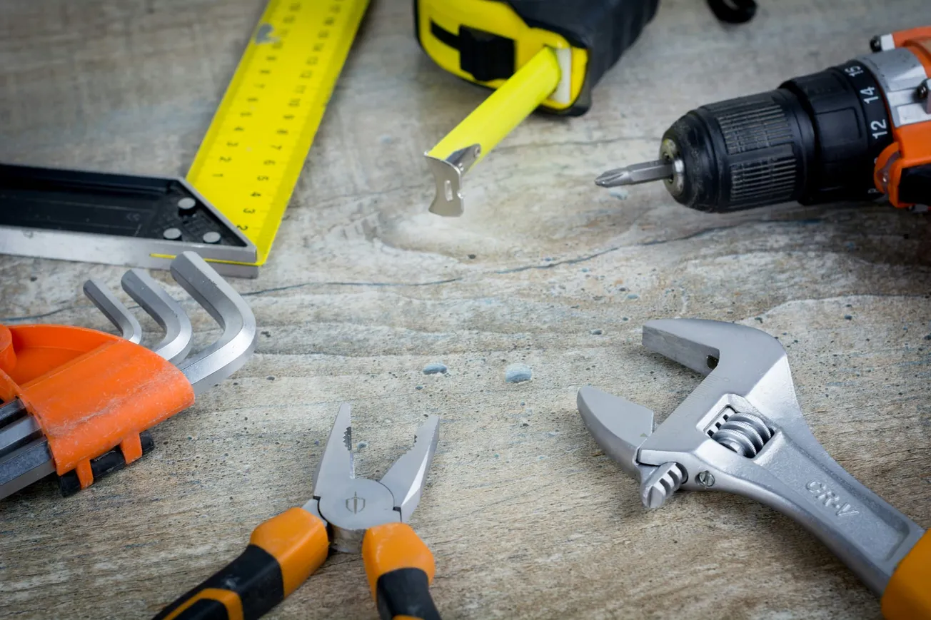 Close-up of various tools on a wooden surface: measuring tape, drill, adjustable wrench, Allen keys, and pliers, conveying a DIY or repair theme.