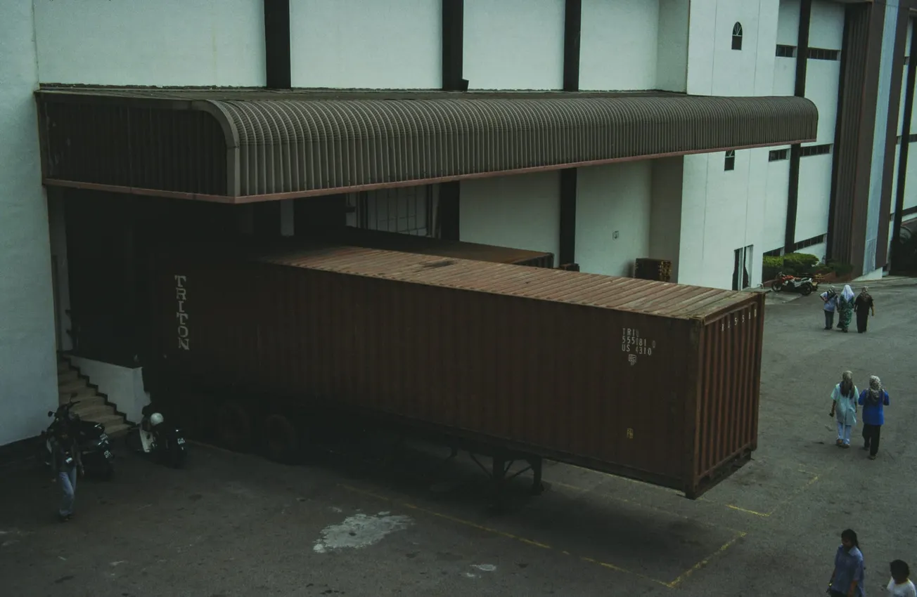 A large shipping container truck is docked at a warehouse loading bay under a corrugated metal roof. Four people are walking nearby. The mood is industrial and busy.