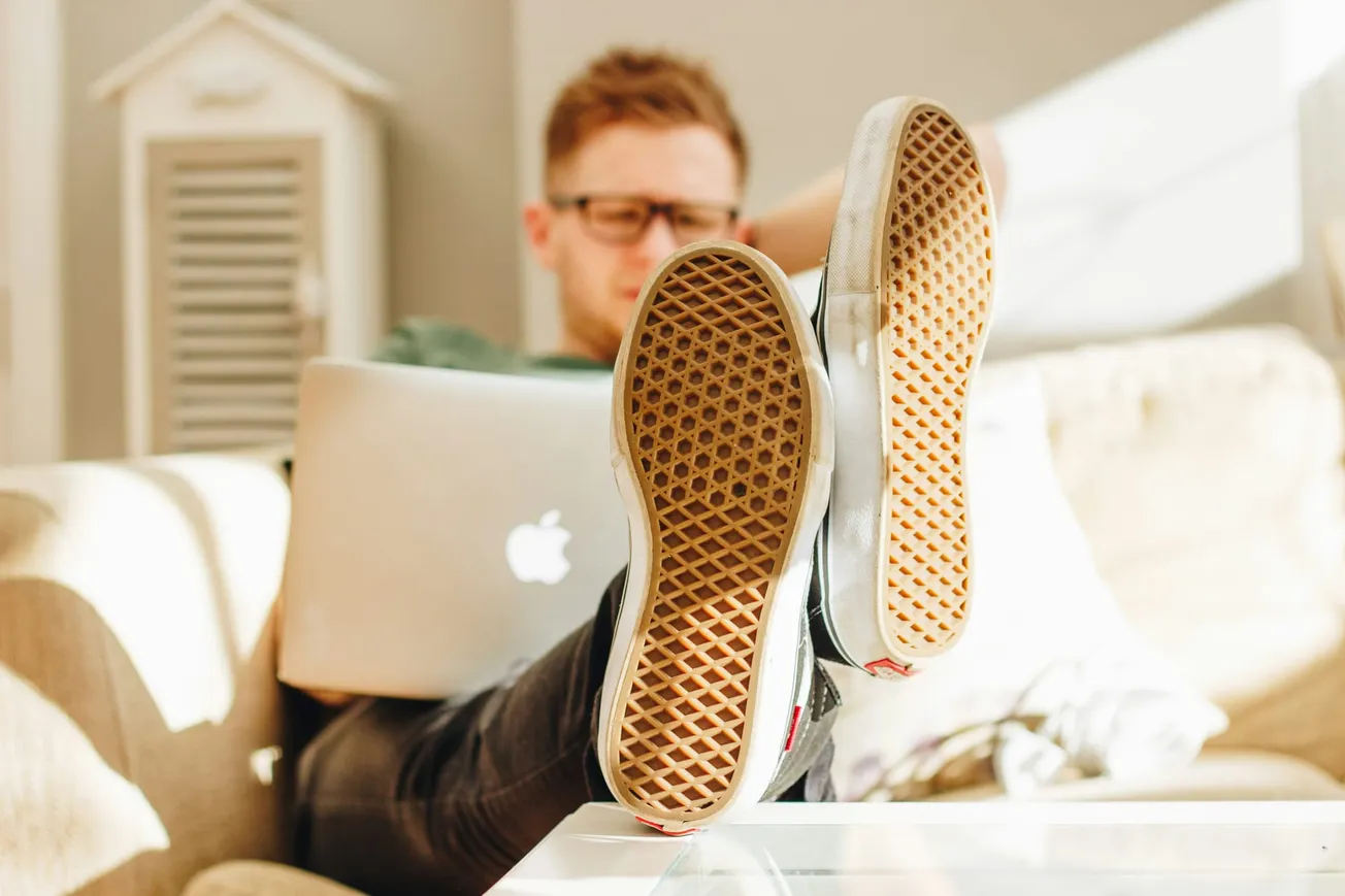 Man in glasses relaxes with feet up on a table, working on a laptop. Sunlight filters in, creating a casual and cozy atmosphere.