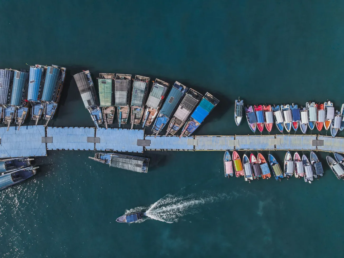 Aerial view of a bustling pier with various boats docked on both sides. A colorful array of boats contrasts with the deep blue water, exuding a lively atmosphere.