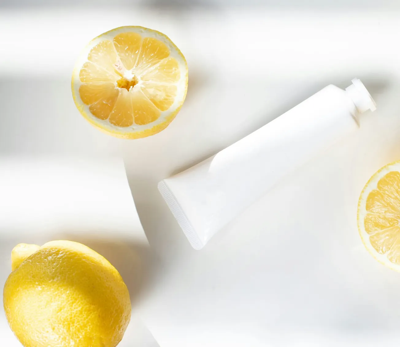 A white tube lies on a bright surface surrounded by halved and whole lemons, creating a fresh, clean, and citrusy atmosphere.