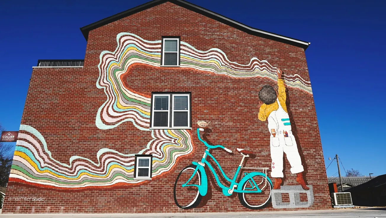 A colorful mural on a brick wall shows a child on a teal bike reaching up, with swirling rainbow lines extending from their hand, conveying creativity and motion.