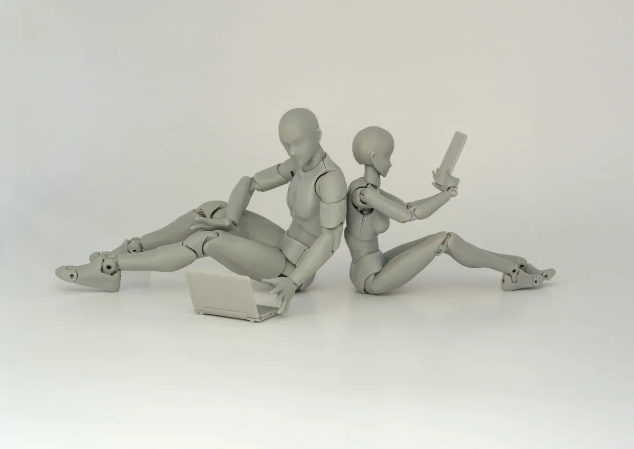 Two gray humanoid figurines sit back-to-back. One uses a laptop, the other holds a smartphone, conveying a theme of technology and communication.