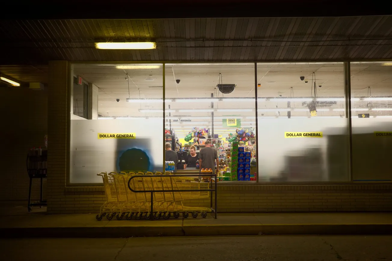 View through a dimly lit dollar store window at night, showing two people inside near colorful shelves. Yellow shopping carts are lined up outside.