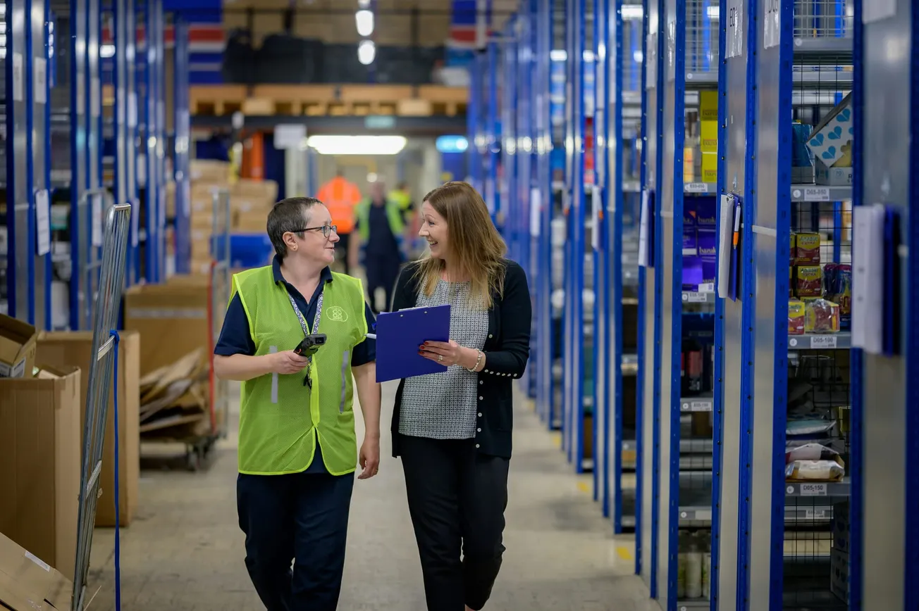 Two people walk through a warehouse aisle, smiling and discussing notes. Shelves are stocked with various items. The scene feels collaborative and busy.