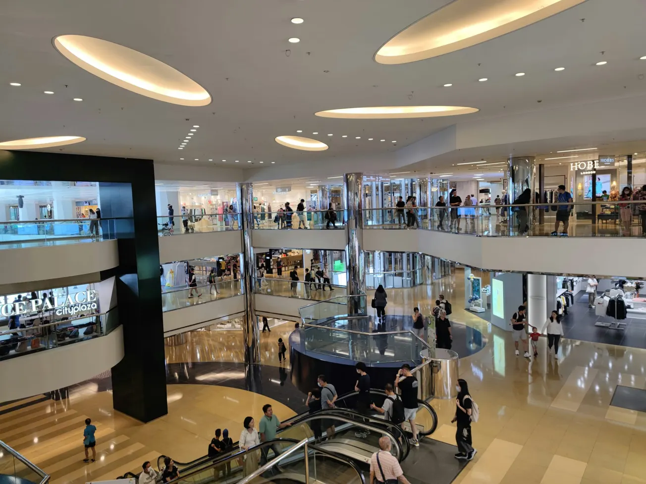 A spacious, multi-level shopping mall interior with sleek modern design. Shoppers fill the floors, and escalators connect levels. Bright, busy ambiance.