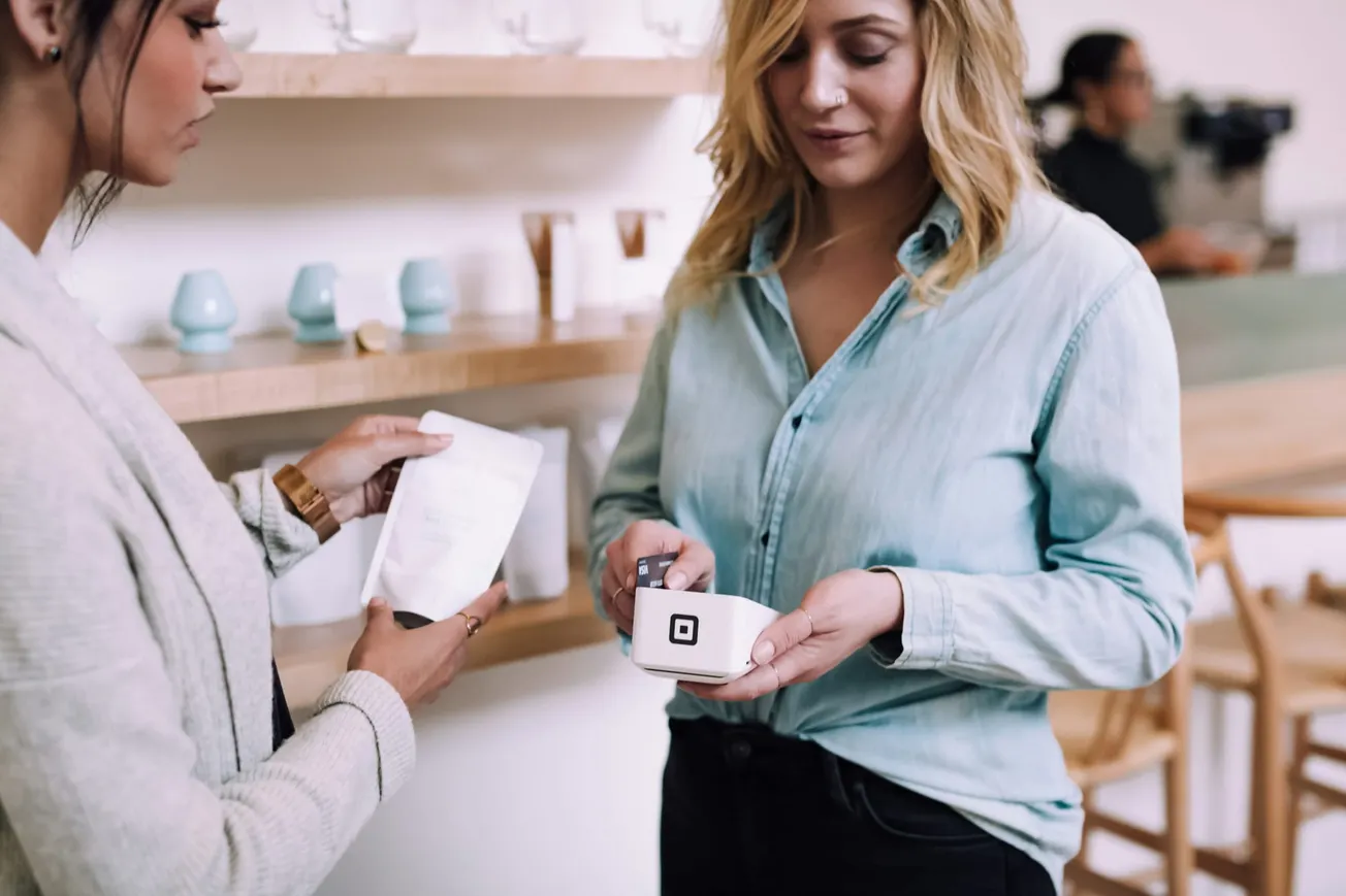 Two women are in a modern cafe. One holds a card reader while processing a payment; the other hands over a product. The atmosphere is calm and focused.