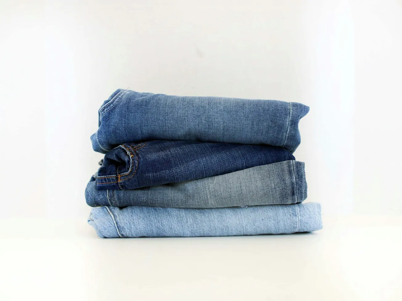 A stack of four neatly folded denim jeans in varying shades of blue sits on a white surface. The background is plain, creating a minimalist look.