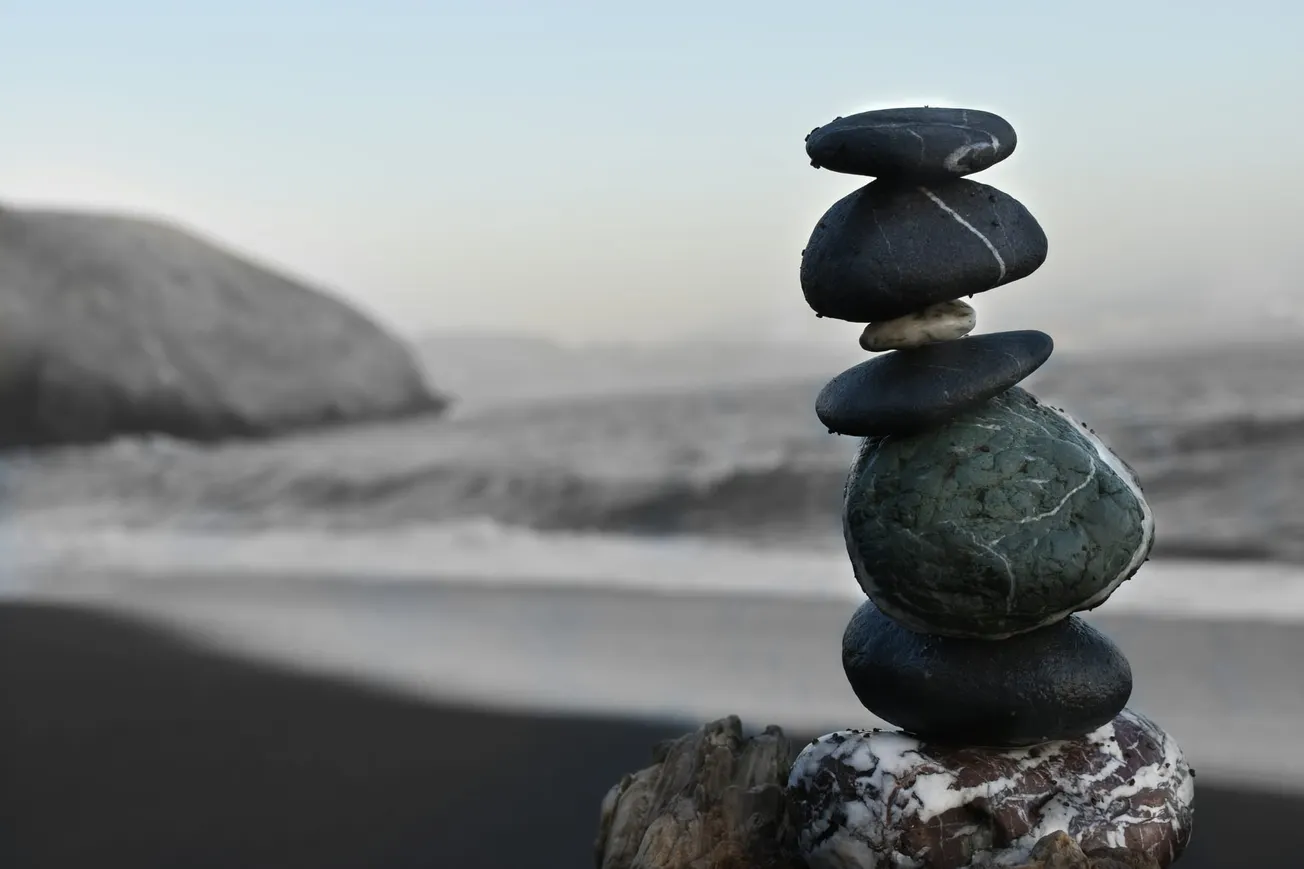 Stacked stones balanced on driftwood on a serene beach, with blurred waves and rocky coast in the background, creating a peaceful, harmonious scene.
