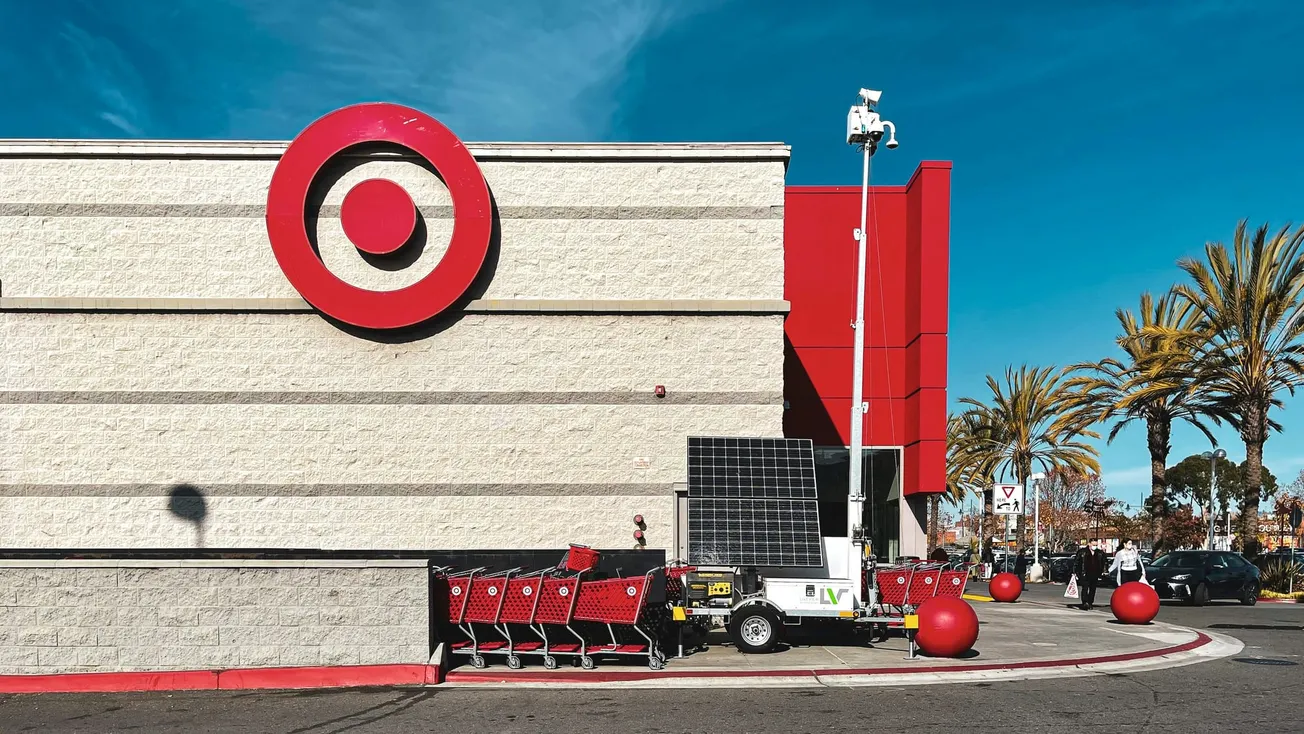 A Target store facade with the large red bullseye logo. Red shopping carts are lined up and a mobile security unit stands nearby. Palm trees and a blue sky in the background.