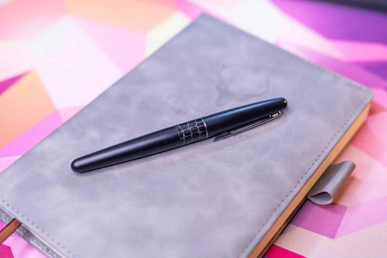 A black pen with a geometric design rests on a gray notebook with a subtle deer logo. The background features a vibrant geometric pattern.