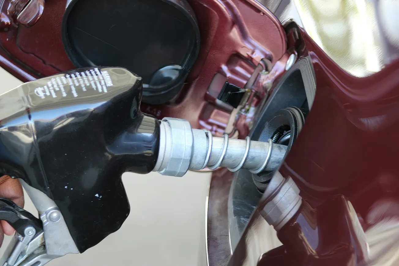 A close-up of a black gas nozzle inserted into a car's fuel tank. The car is maroon, and the shiny nozzle reflects light, conveying utility and focus.