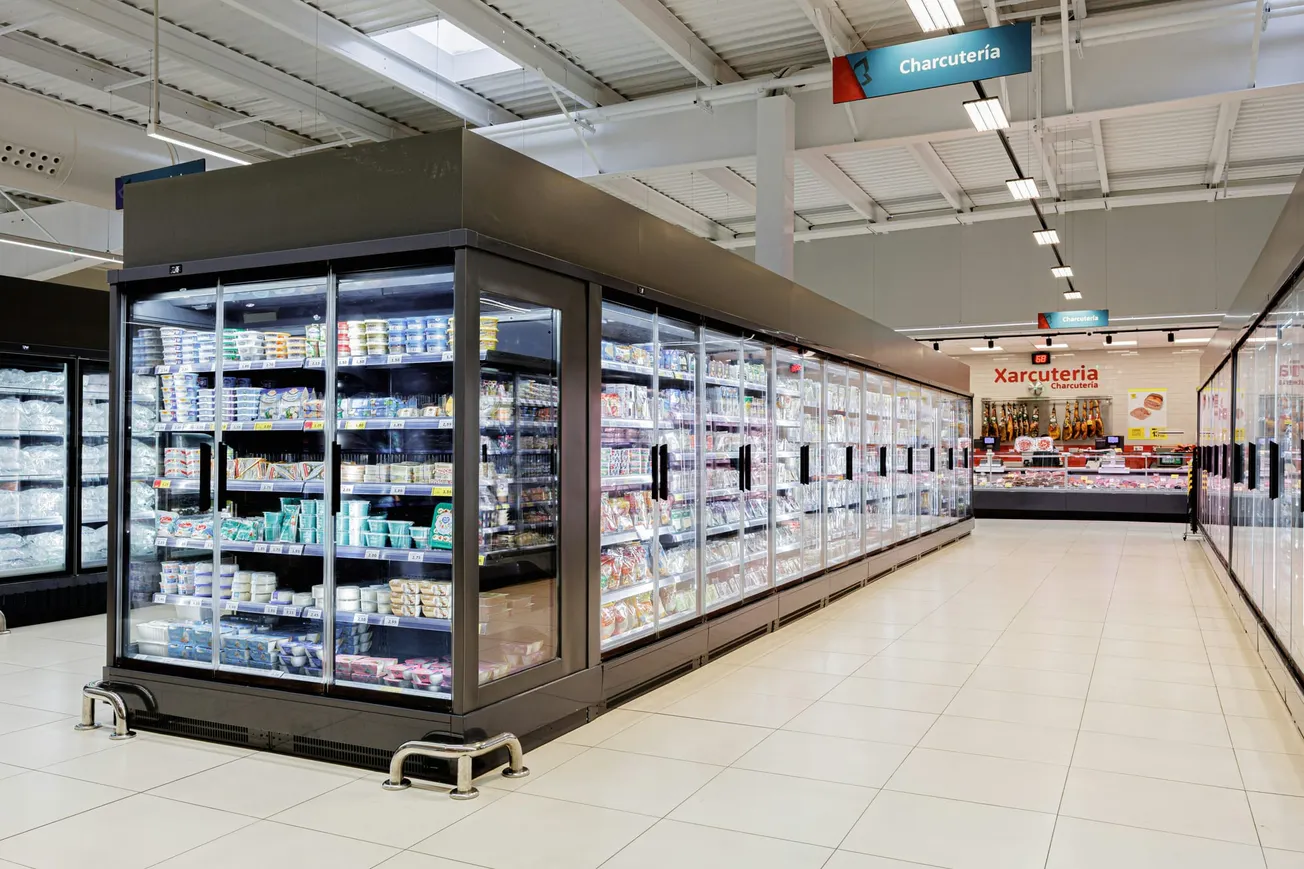 Spacious supermarket aisle with illuminated refrigerated shelves displaying various dairy products. Overhead signs in Spanish indicate the deli section.