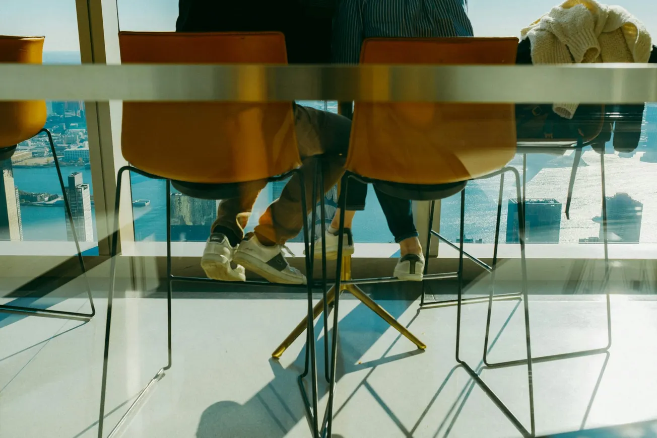 Two people sit at a glass-top table by a window with a city and water view. Orange chairs and sunlight create a warm, relaxed atmosphere.