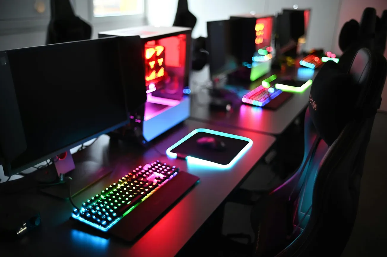 A gaming setup with glowing RGB keyboards, mice, and LED-lit PCs on a black desk. The mood is vibrant and futuristic, with a focus on technology.