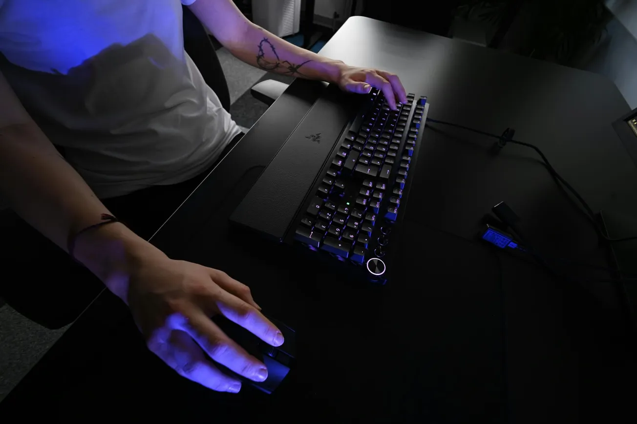 A person with tattoos on their forearm uses a gaming keyboard and mouse in a dimly lit room. Blue light illuminates their hands, creating a focused atmosphere.