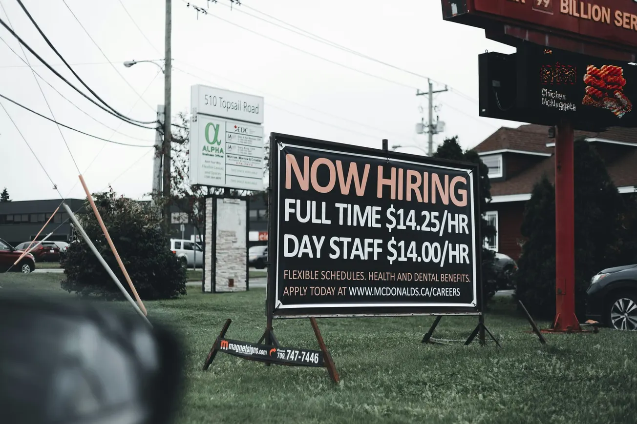 Outdoor sign at a roadside advertises job openings. It reads, "Now Hiring: Full Time $14.25/HR, Day Staff $14.00/HR," against a backdrop of buildings and cars.