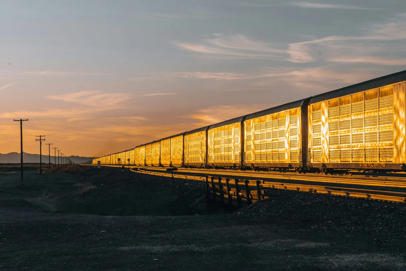 Freight train cars illuminated in golden light at sunset, stretching into the distance. The sky is warm with streaks of cloud, and telephone poles line the tracks.