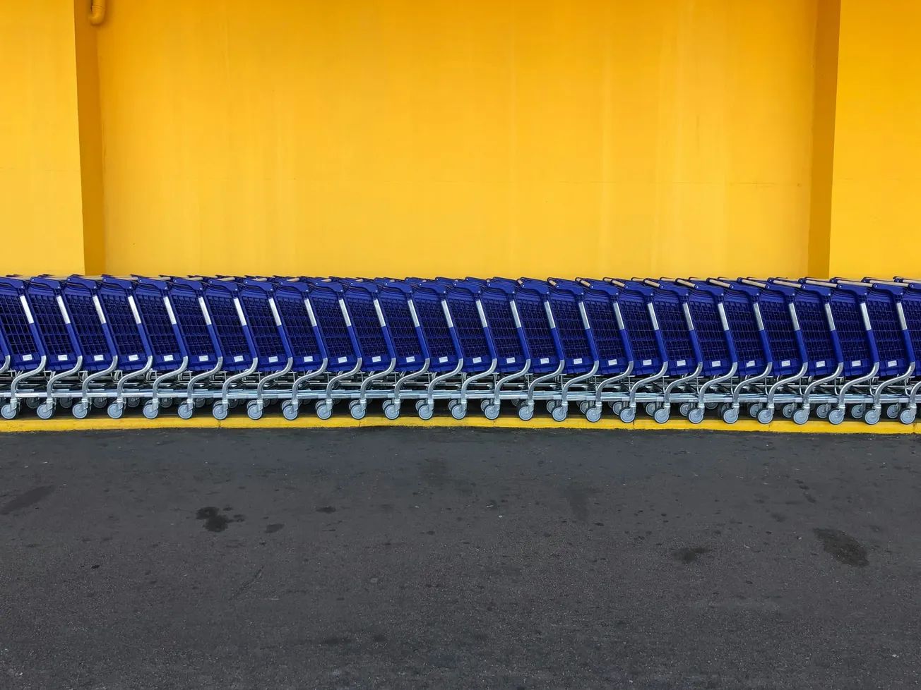 A neat row of blue shopping carts is aligned in front of a bright yellow wall, creating a striking color contrast and an orderly, geometric pattern.