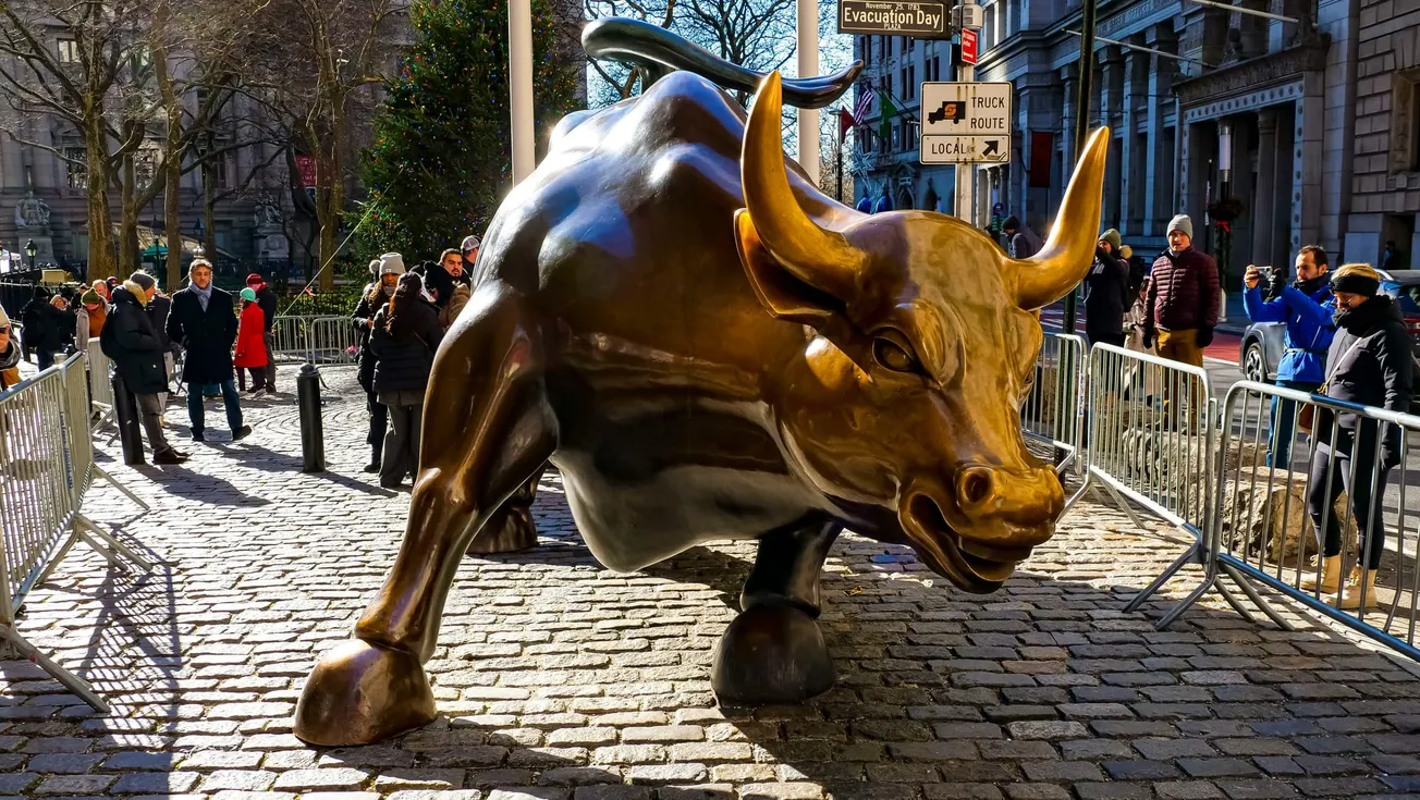 A large bronze bull statue on a cobblestone street surrounded by metal barriers. People are gathered around, taking photos. The mood is lively and vibrant.