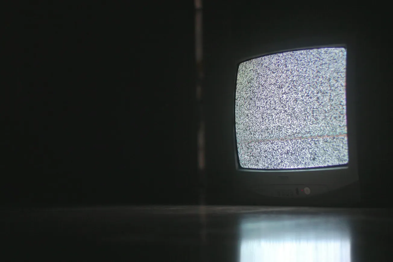 A TV in a dark room displays static, creating an eerie, nostalgic atmosphere. The screen’s light reflects on the floor, emphasizing isolation.