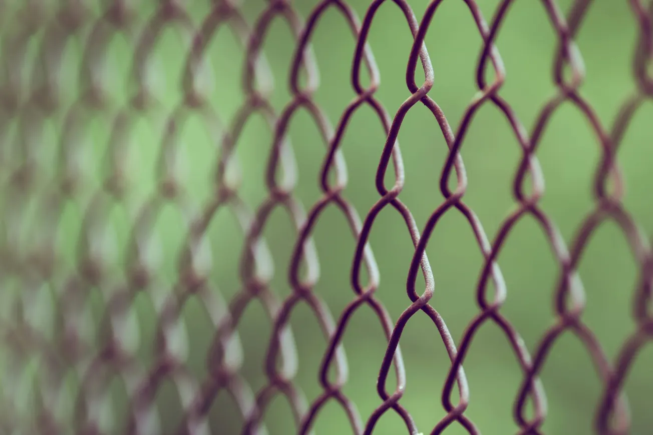Close-up of a rusty chain-link fence, with diamond shapes, against a blurred green background, conveying a sense of isolation and simplicity.