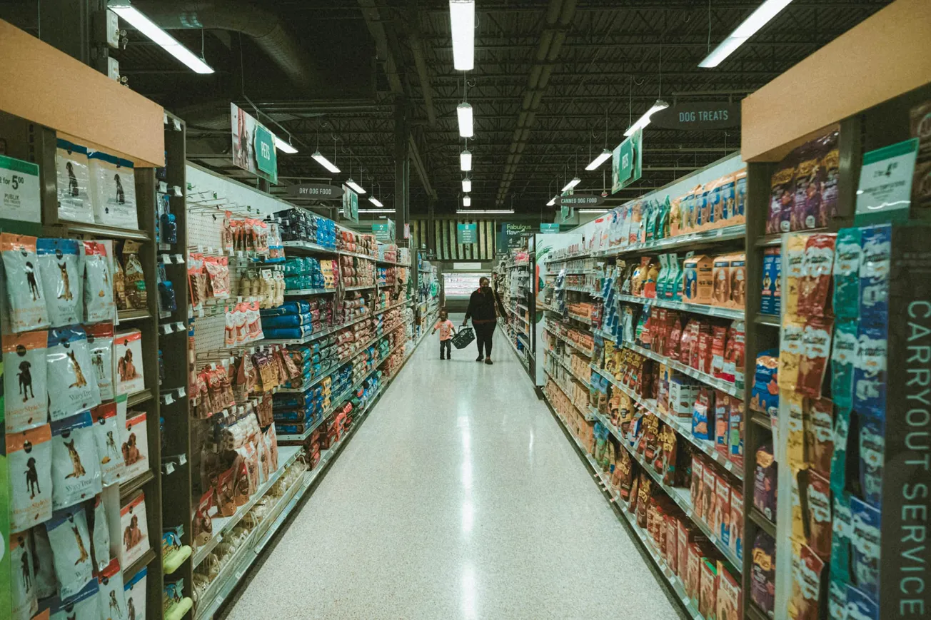 Wide supermarket aisle with pet food on both sides. A person and child walk down the brightly lit aisle, creating a calm, everyday shopping scene.