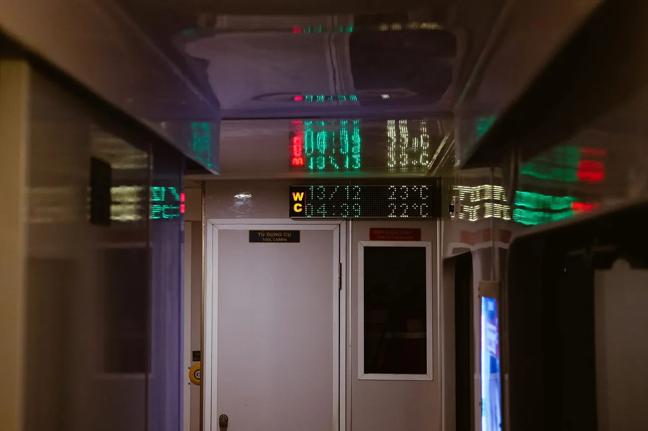 Dimly lit train corridor with a glowing "WC" sign above a white door. Reflections of green and red digital text illuminate the ceiling.