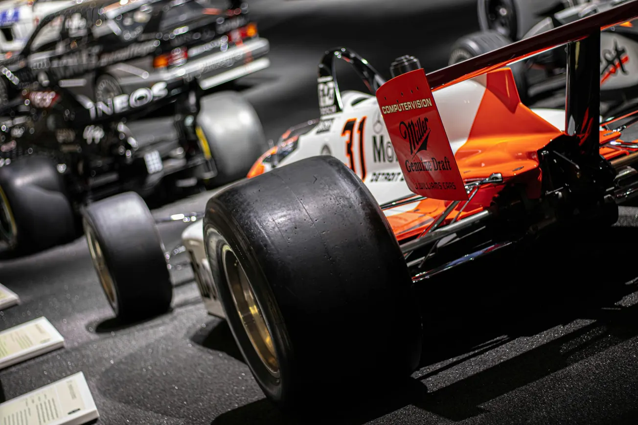 Close-up of a vintage Formula 1 car displaying bold orange and white colors, large rear tires, and sponsor logos, with other race cars blurred in the background.