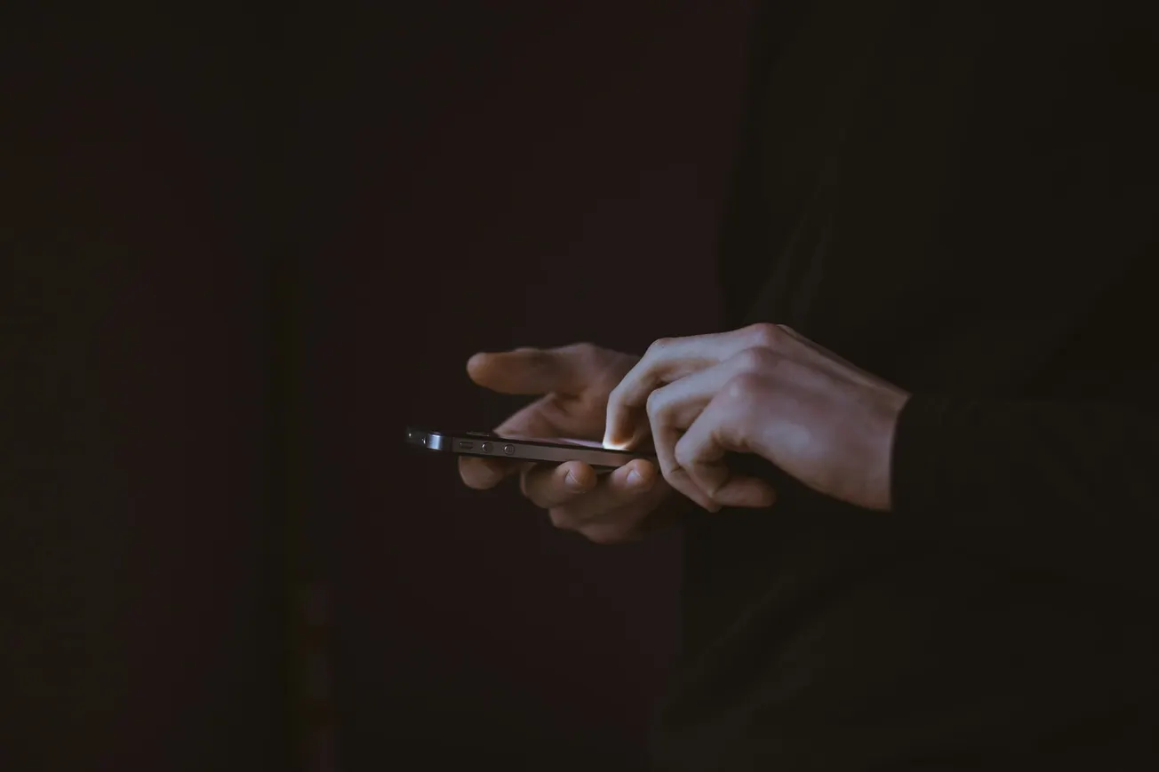 A person in a dark sweater uses a smartphone in low light, focusing intensely. The dim background and soft lighting create a calm and introspective mood.