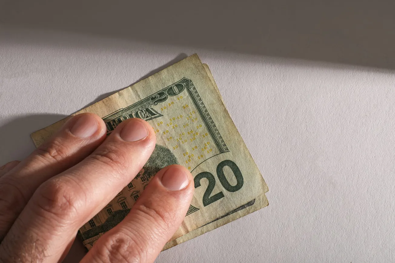 A hand holding a folded twenty-dollar bill on a gray surface. The lighting casts a soft shadow, creating a sense of secrecy or discretion.