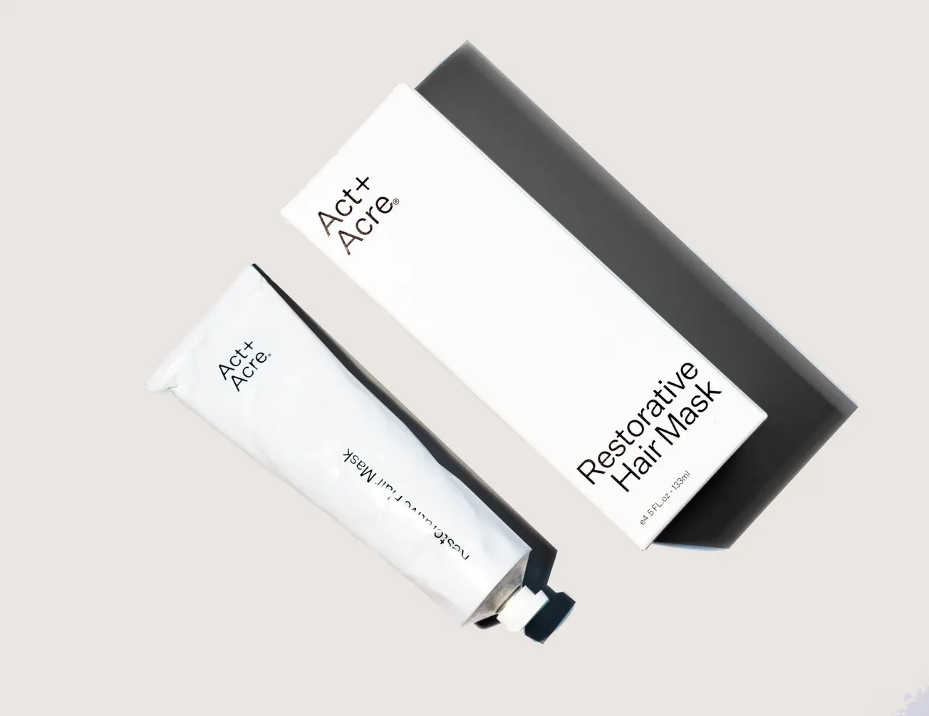 A white Act+Acre Restorative Hair Mask tube lies next to its sleek black and white box, conveying a minimalist and sophisticated feel.