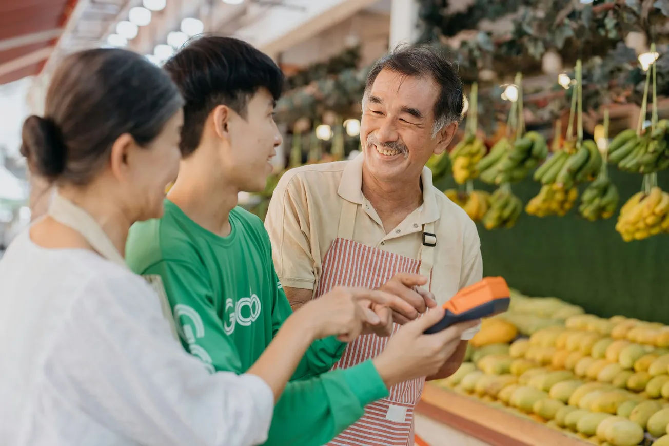 A smiling fruit vendor in an apron interacts warmly with two customers in a market. They hold a payment device amid vibrant hanging bananas.