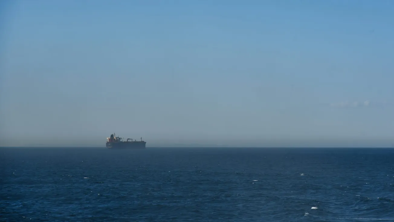 A large cargo ship sits on the horizon of a vast, deep blue ocean under a clear, hazy sky.