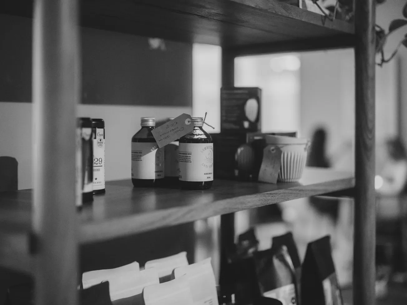 A black and white, shallow-focus photograph of apothecary-style glass bottles and various packaged goods arranged on a wooden shelf.