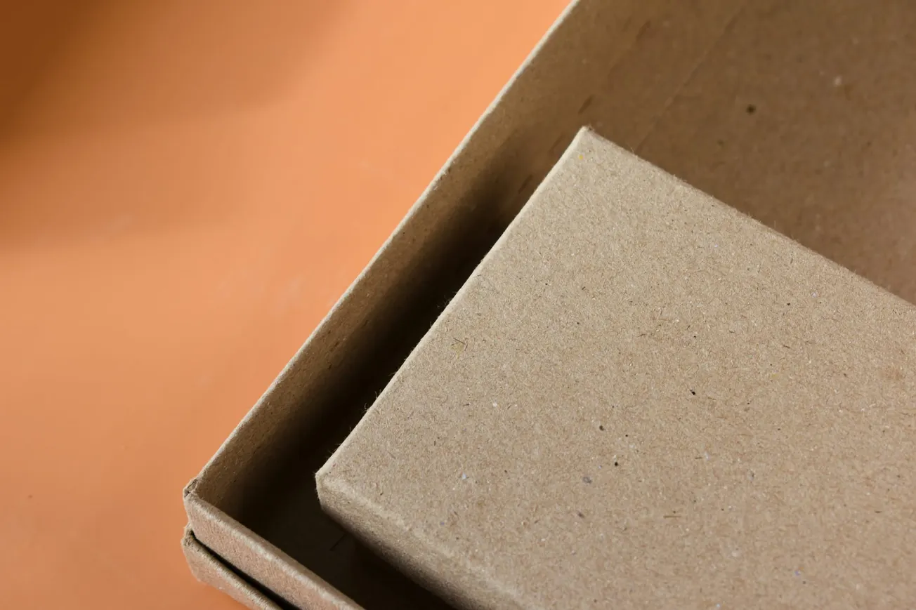 Close-up of an open cardboard box against an orange background. The box is empty, highlighting its textured, brown surface and angular edges.