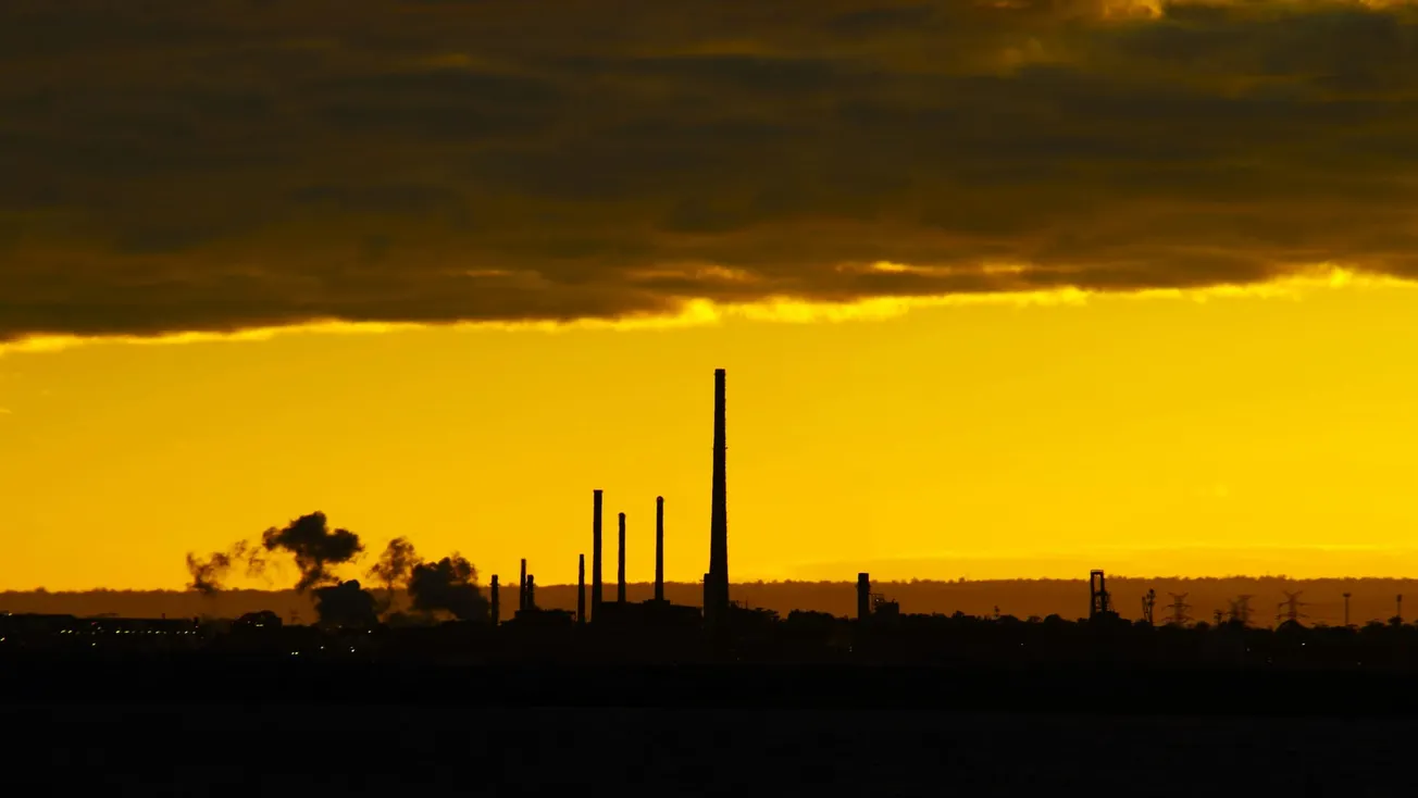 Industrial landscape at sunset, with tall smokestacks silhouetted against a glowing yellow sky. Dark clouds hover above, creating a dramatic contrast.