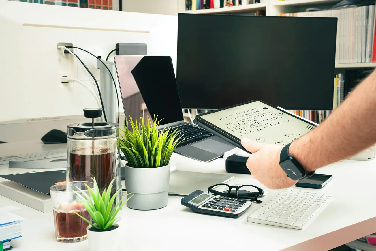 A cluttered desk with plants, a calculator, coffee, computers, and a person holding a tablet with sheet music. The scene feels busy yet organized.
