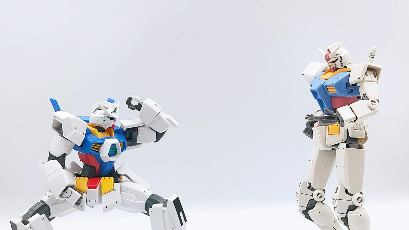 Two Gundam models are set against a plain white backdrop. The left figure is crouched with an arm raised, while the right stands upright. Both have vivid blue, red, and yellow accents.