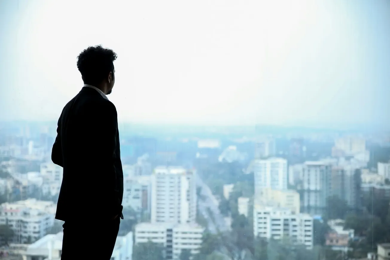 A man in a suit stands gazing out a large window overlooking a cityscape with tall buildings. The scene conveys a sense of contemplation and ambition.