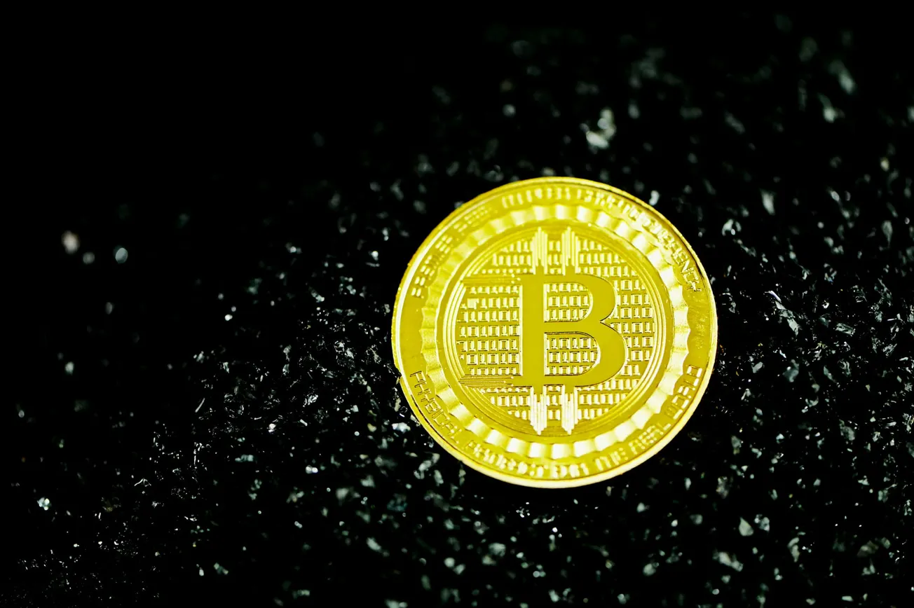 A shiny gold Bitcoin coin on a textured black surface. The coin's intricate details and the prominent Bitcoin symbol suggest wealth and digital finance.