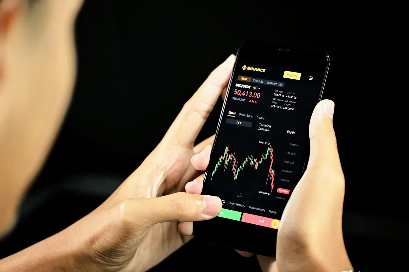 A person holds a smartphone displaying a cryptocurrency trading app. The screen shows Bitcoin's price chart with real-time data and a dark background.