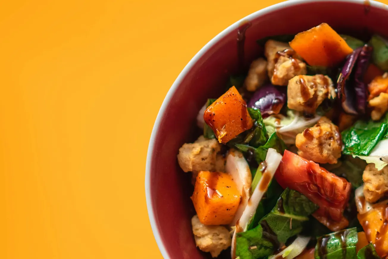 A vibrant salad in a red bowl against an orange background features roasted squash, leafy greens, tofu pieces, and red cabbage, creating a fresh and colorful dish.