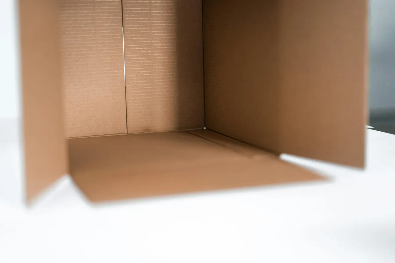 Open brown cardboard box on a white surface, flaps extended. The box is empty, showcasing textural details and clean lines, giving a minimalist feel.