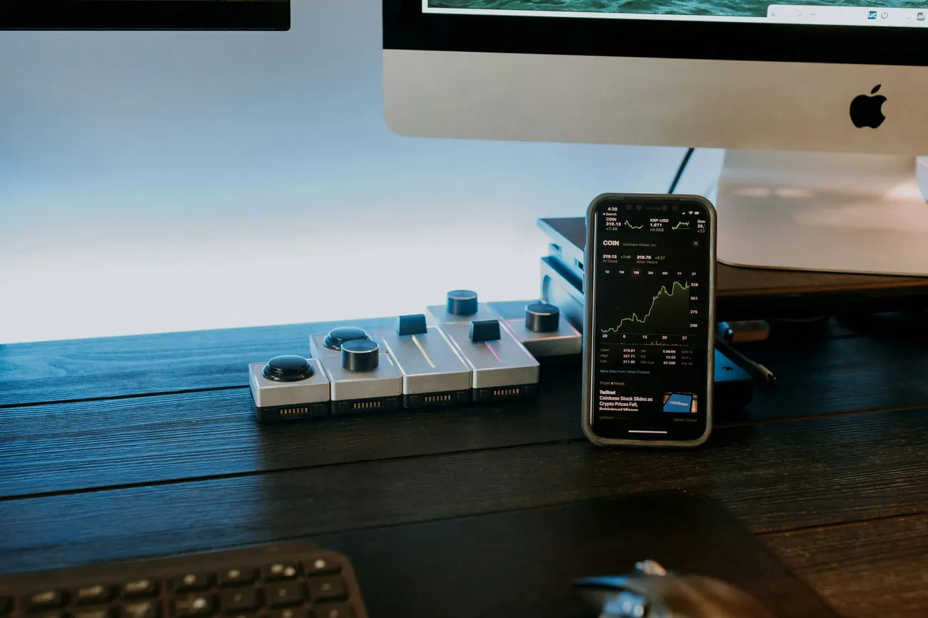 A sleek workspace featuring a smartphone displaying stock market graphs, set against a desktop computer on a dark wooden surface. Tech-savvy and modern ambiance.