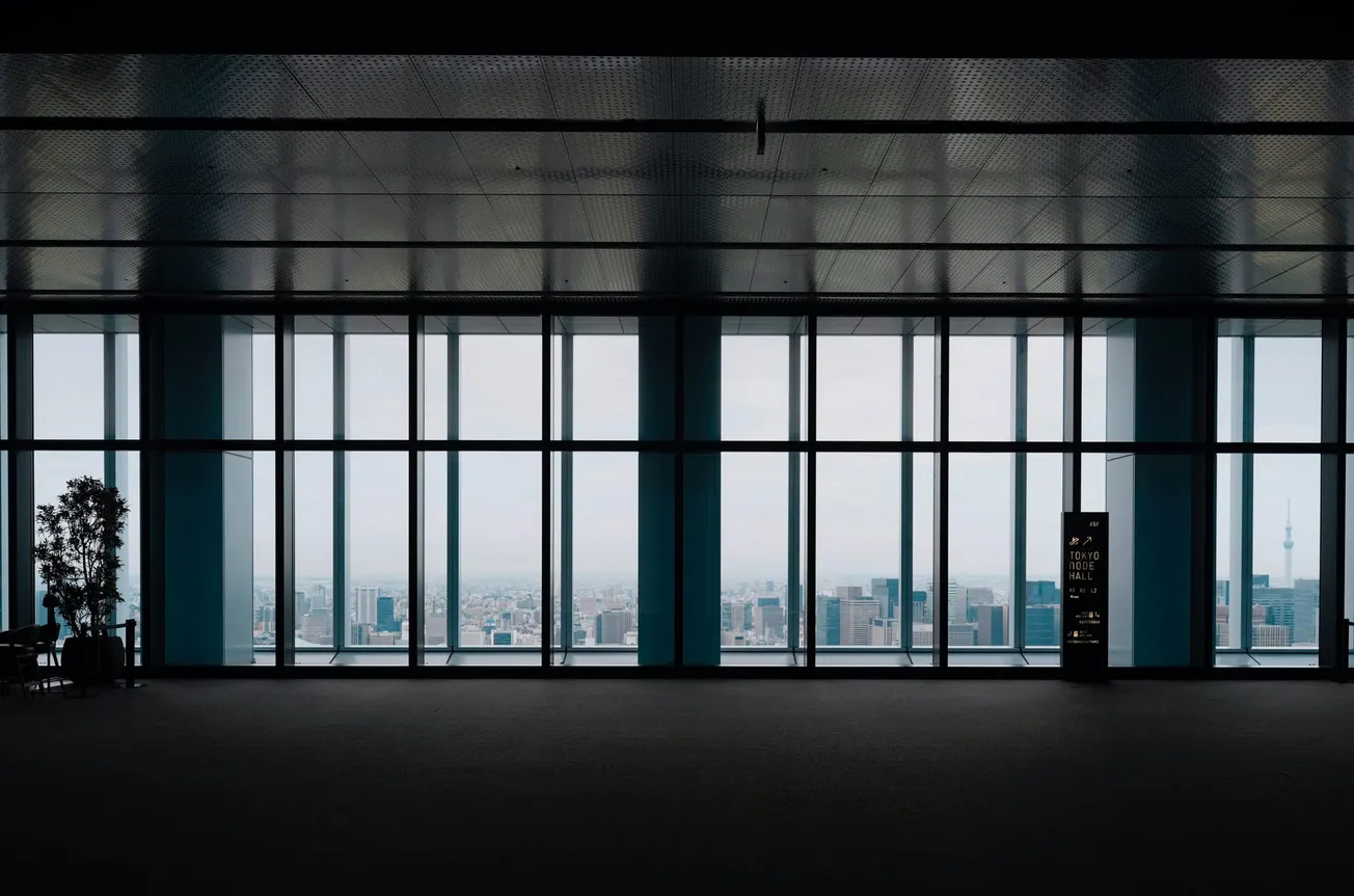 A large, empty room with floor-to-ceiling windows offers a panoramic city view. A lone potted plant and a vending machine are silhouetted, creating a serene mood.